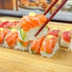 A hand using chopsticks to pick up a piece of sushi, topped with raw salmon, avocado, and sesame seeds, from a row of sushi pieces on a wooden board.