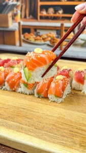 A close-up of chopsticks holding a piece of Ice Cube Tray Sushi topped with salmon, avocado, and a small dollop of sauce, above a wooden board with more sushi rolls in the background.