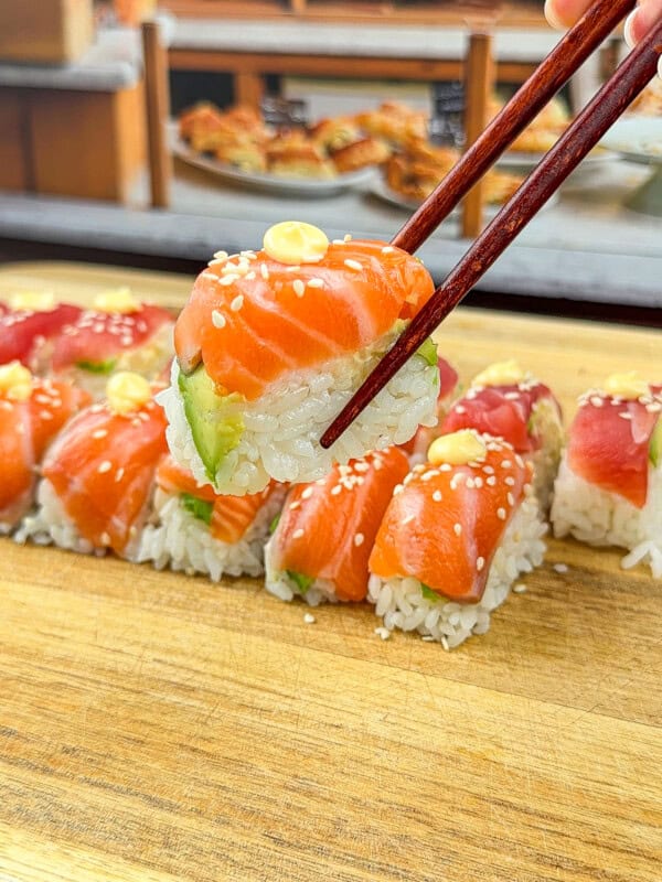 A close-up of chopsticks holding a piece of Ice Cube Tray Sushi topped with salmon, avocado, and a small dollop of sauce, above a wooden board with more sushi rolls in the background.