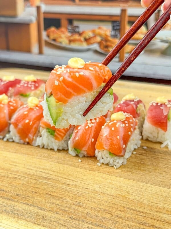 A hand using chopsticks to pick up a piece of sushi, topped with raw salmon, avocado, and sesame seeds, from a row of sushi pieces on a wooden board.