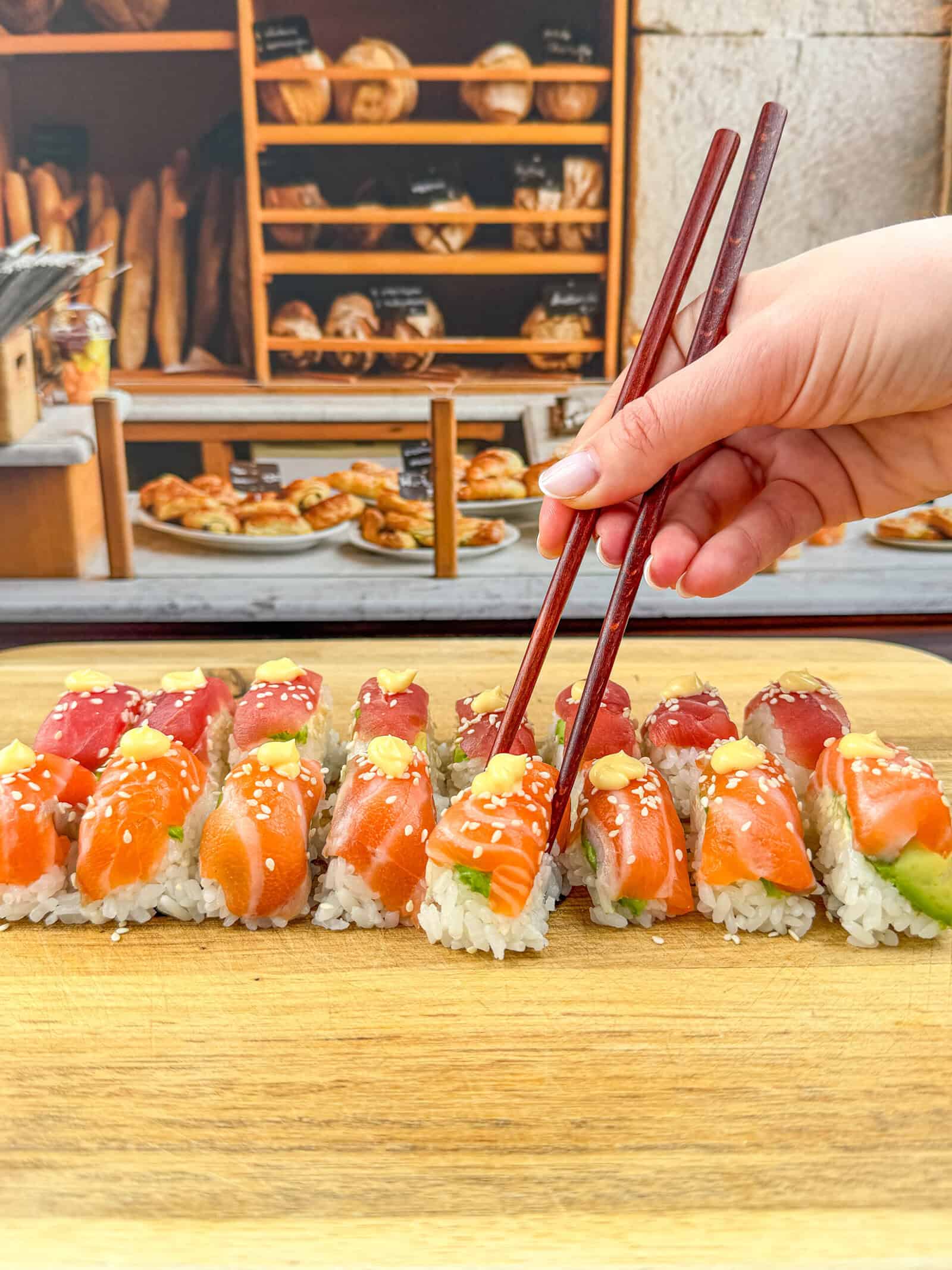 A hand holding chopsticks picks up a piece of sushi from a wooden board topped with assorted nigiri; shelves of bread are visible in the background.