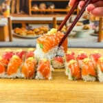 A hand holding chopsticks lifts a piece of Ice Cube Tray Sushi—salmon topped with sesame seeds—from a wooden board lined with more sushi rolls; bakery shelves are blurred in the background.