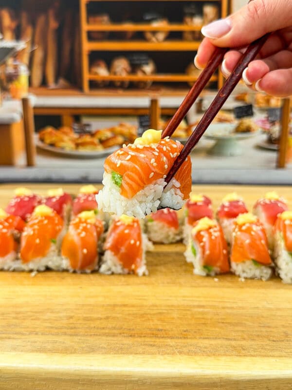 A hand holding chopsticks lifts a piece of Ice Cube Tray Sushi—salmon topped with sesame seeds—from a wooden board lined with more sushi rolls; bakery shelves are blurred in the background.