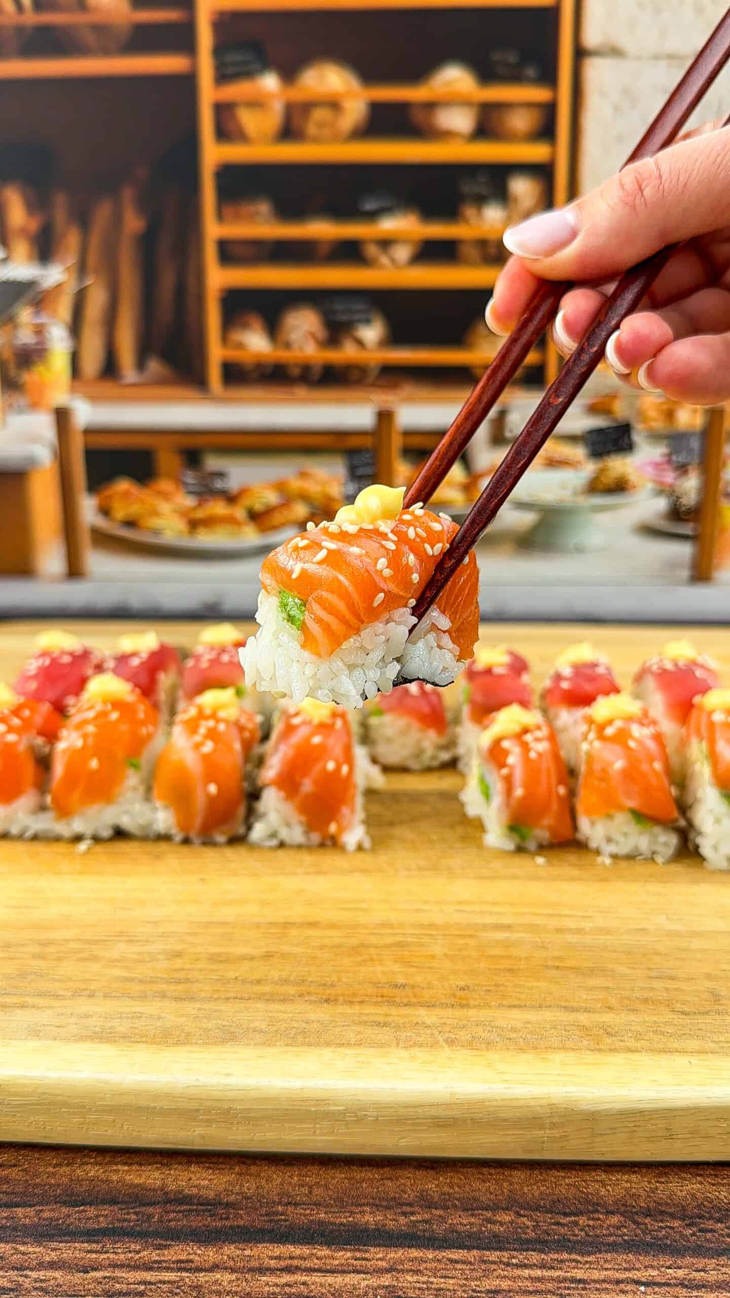 A hand holding chopsticks lifts a piece of Ice Cube Tray Sushi—salmon topped with sesame seeds—from a wooden board lined with more sushi rolls; bakery shelves are blurred in the background. - 3