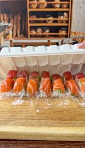 An ice cube tray is being used to shape sushi pieces wrapped in plastic wrap, placed on a wooden surface. A bakery shelf with bread loaves is visible in the background.