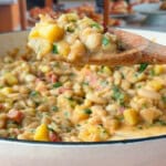 A wooden spoon holds a scoop of creamy bean and potato stew over a white pot, with visible chunks of yellow squash, beans, and herbs. In the blurred background, shelves display various breads and baked goods.