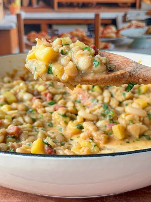 A wooden spoon holds a scoop of creamy bean and potato stew over a white pot, with visible chunks of yellow squash, beans, and herbs. In the blurred background, shelves display various breads and baked goods.