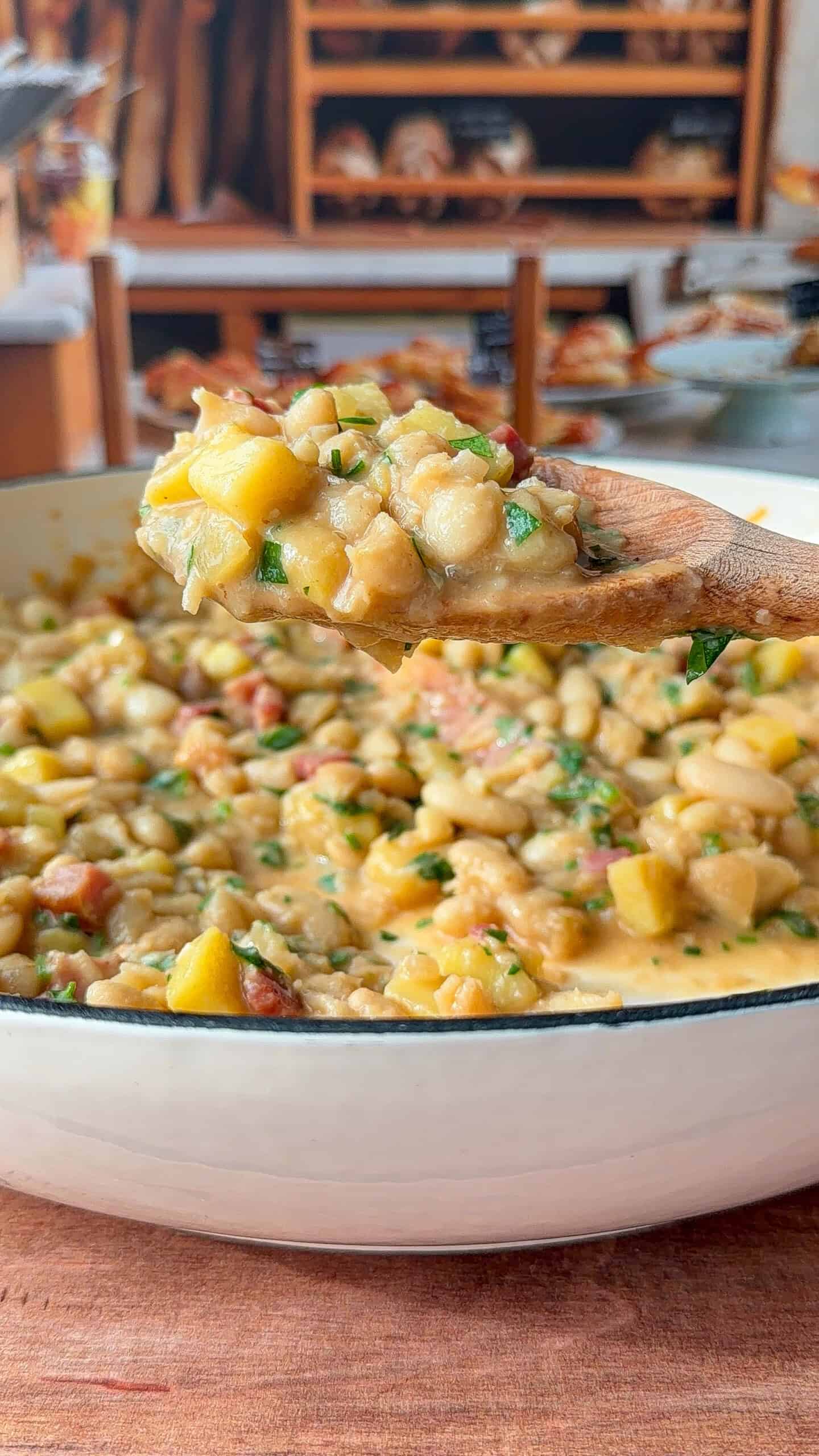 A wooden spoon holds a scoop of creamy bean and potato stew over a white pot, with visible chunks of yellow squash, beans, and herbs. In the blurred background, shelves display various breads and baked goods.