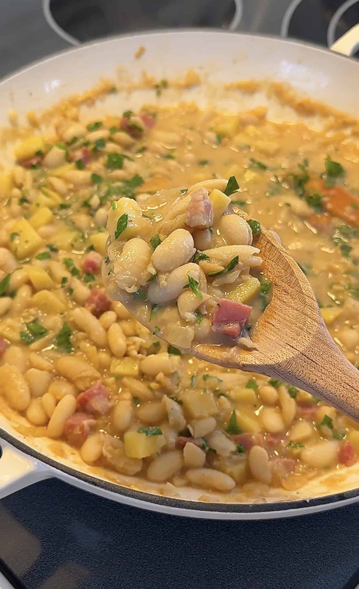 A wooden spoon holds up a serving of Creamy Bean and Potato Stew with chopped herbs and diced ham over a pot filled with the same creamy mixture.