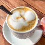 A hand holds a cup of viral egg coffee topped with frothy milk foam and a metal spoon, placed on a white saucer on a wooden table in bright sunlight.