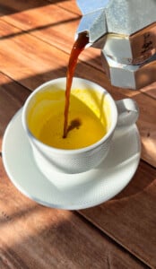 Coffee is being poured from a moka pot into a white cup containing a yellow frothy viral egg coffee mixture, set on a matching saucer on a wooden table bathed in sunlight and shadows.