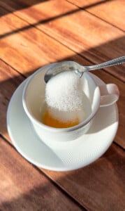 A spoonful of granulated sugar is being added to a white cup of viral egg coffee on a matching saucer, set on a wooden table with sunlight casting shadows.