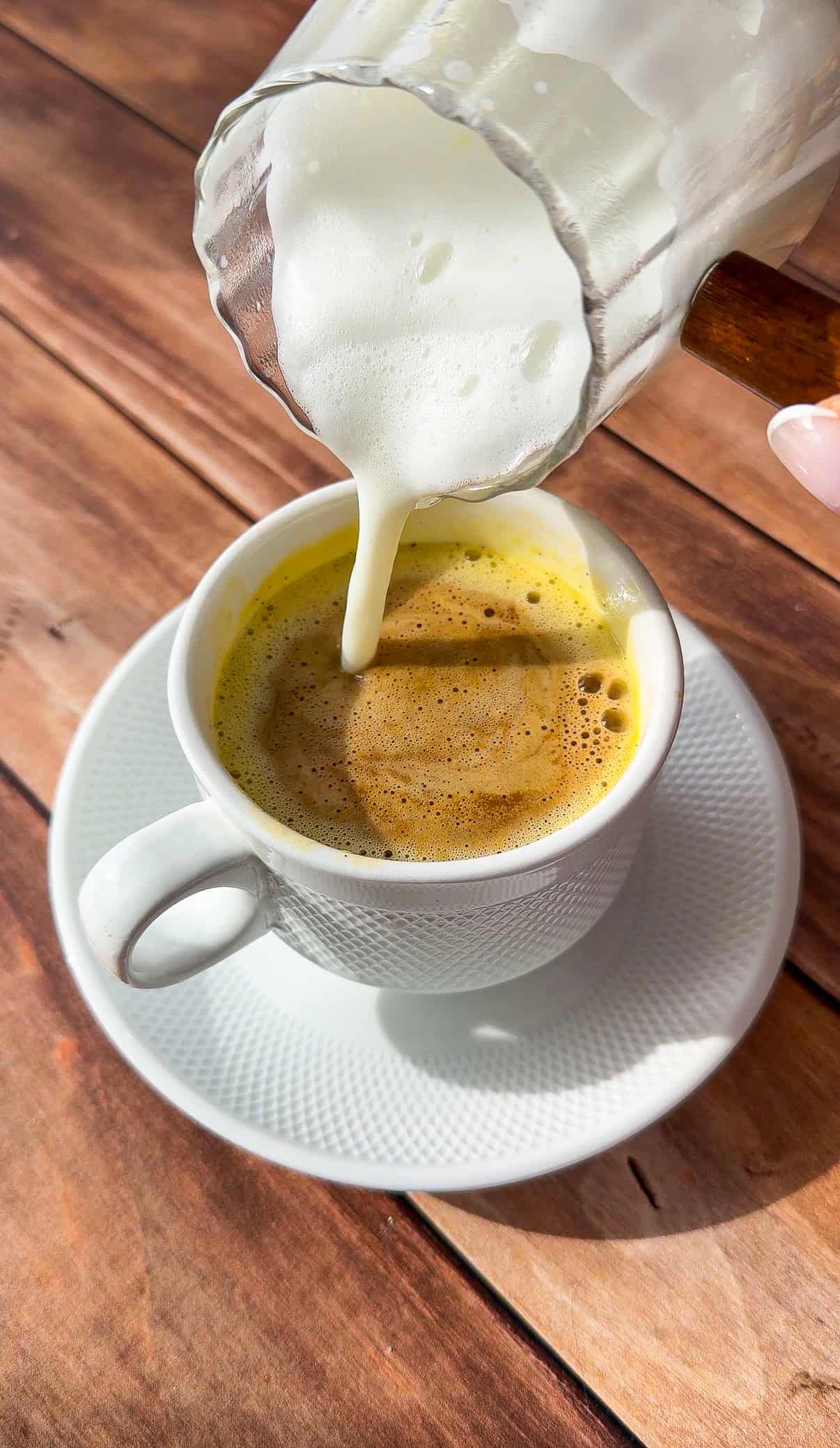 A hand pours steamed milk from a glass pitcher into a cup of viral egg coffee on a white saucer, set on a wooden table in natural sunlight.