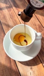 A stream of brown liquid is being poured into a white cup on a saucer, containing egg yolks and condensed milk—creating viral egg coffee—on a wooden table bathed in sunlight.