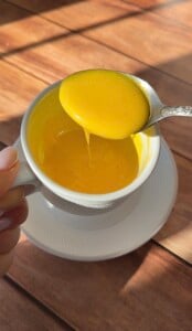 A hand holds a white cup filled with thick, bright yellow viral egg coffee while a spoon lifts some of the creamy liquid, letting it slowly drip back into the cup. The cup rests on a matching saucer atop a wooden surface.