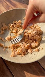 A hand uses a fork to shred cooked salmon in a speckled beige bowl on a wooden surface.