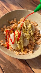 A bowl with shredded salmon and two rice balls, drizzled with yellow mayonnaise and red sriracha sauce, sits on a wooden surface. A green squeeze bottle dispenses sriracha over the dish.