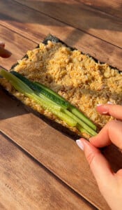 Close-up of hands rolling sushi with a sheet of seaweed, a layer of rice and tuna mixture, and strips of cucumber on a wooden surface. Sunlight casts a warm glow on the scene.