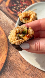 A hand with light gray nail polish holds a piece of sushi wrapped in seaweed with brown rice, vegetables, and sauce, over a white plate on a wood-textured surface.