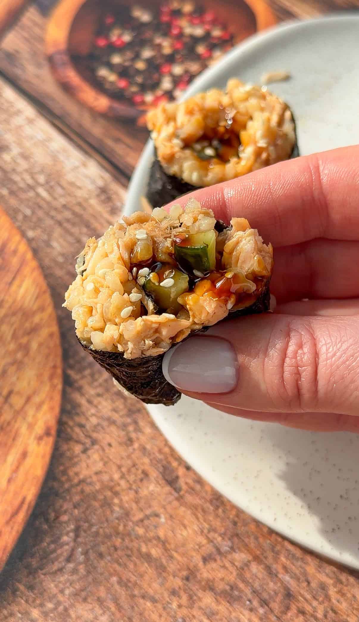 A hand with light gray nail polish holds a piece of sushi wrapped in seaweed with brown rice, vegetables, and sauce, over a white plate on a wood-textured surface.