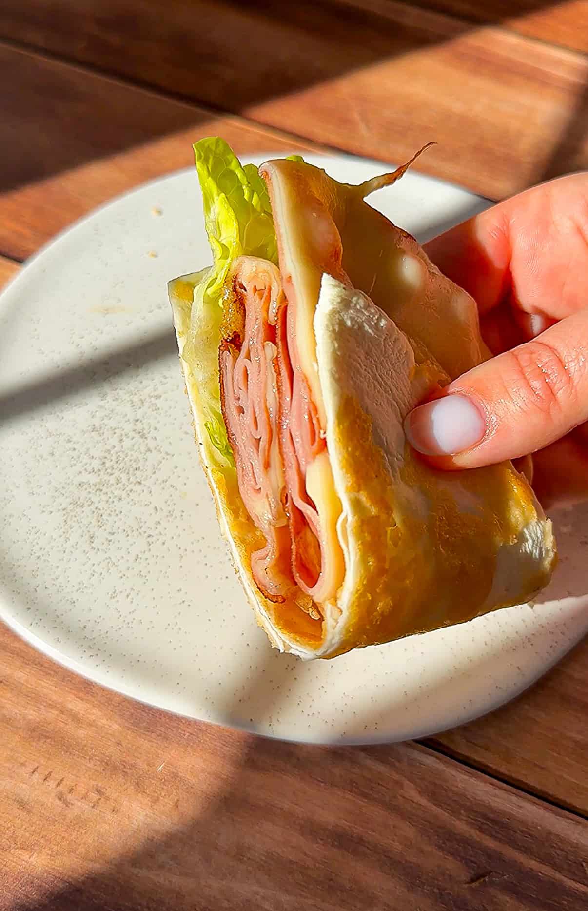 A hand holding a folded wrap filled with Healthy Chicken Tenders, cheese, and lettuce over a white plate on a wooden table in sunlight.