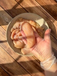 A hand wearing a disposable glove holds a raw chicken breast over a bowl of sliced pieces, ready to make healthy chicken tenders. Sunlight streams across the wooden surface, highlighting the fresh preparation.