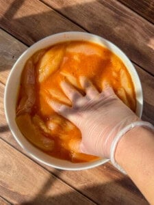 A gloved hand mixes Healthy Chicken Tenders in a white bowl filled with orange marinade, placed on a wooden surface in sunlight.