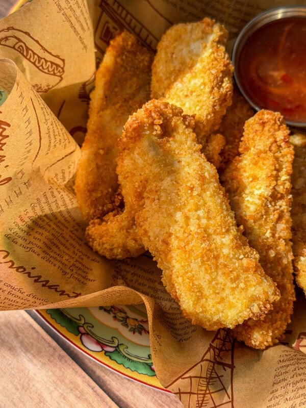 Golden, crispy Healthy Chicken Tenders served in a basket lined with printed paper, with a small metal cup of red dipping sauce on the side.