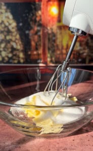 A close-up of a hand mixer blending butter and sugar in a clear glass bowl on a wooden surface, with a blurred, warm-toned background.