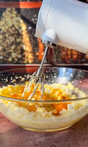 A hand mixer blends eggs and sugar in a glass bowl, with daylight streaming through a window in the background.