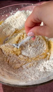 A hand holding a fork mixes flour into a yellowish dough in a glass bowl, combining the ingredients to form batter.