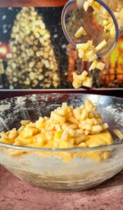 A close-up of chopped apples being poured from a small container into a glass mixing bowl with dough. In the blurred background, a lit Christmas tree and festive lights are visible.