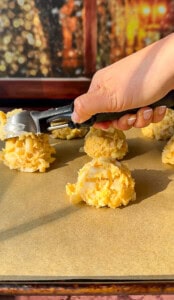 A hand uses a cookie scoop to place rounded scoops of unbaked dough onto a parchment-lined baking sheet, with more dough portions visible in the background.