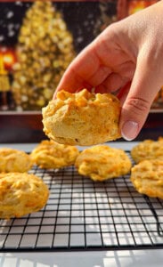 A hand holding a freshly baked biscuit above a cooling rack with several more biscuits, with a blurry, festive background featuring warm lights and a Christmas tree.