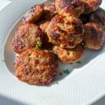 A white plate filled with several golden-brown, pan-fried meatballs, garnished with chopped chives, sits on a white surface in bright natural light.