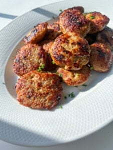 A white plate filled with golden-brown, pan-fried meat patties garnished with chopped fresh chives, shown in bright natural light.