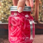 A hand holds a mason jar filled with bright pink pickled onions in brine, with a sprig of dill visible inside. The background is blurred, featuring warm, bokeh lights.
