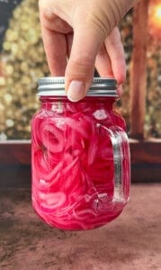 A hand holds a mason jar filled with bright pink pickled onions in brine, with a sprig of dill visible inside. The background is blurred, featuring warm, bokeh lights.