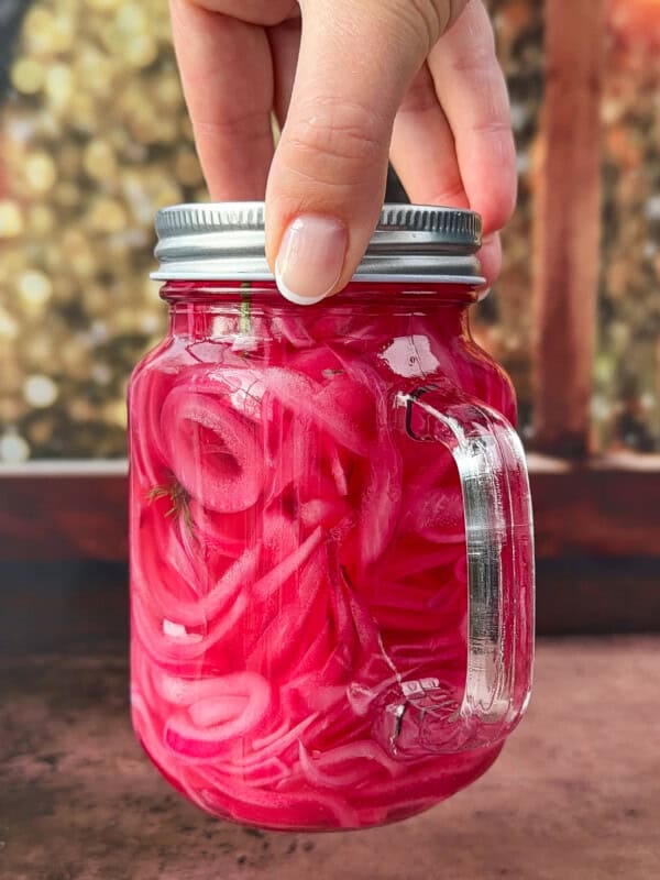 A hand holds a mason jar filled with bright pink pickled onions in brine, with a sprig of dill visible inside. The background is blurred, featuring warm, bokeh lights. - 8