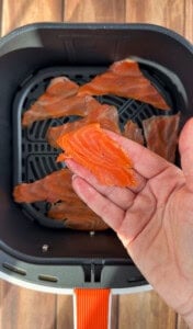 A hand holds a slice of raw salmon over an air fryer basket filled with several other slices of raw salmon, ready to be cooked. The air fryer has a black interior and an orange handle.