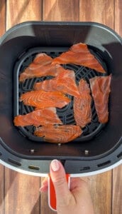 Thin slices of raw salmon are arranged in a single layer inside an air fryer basket, held by a hand with light pink nail polish against a wooden background.