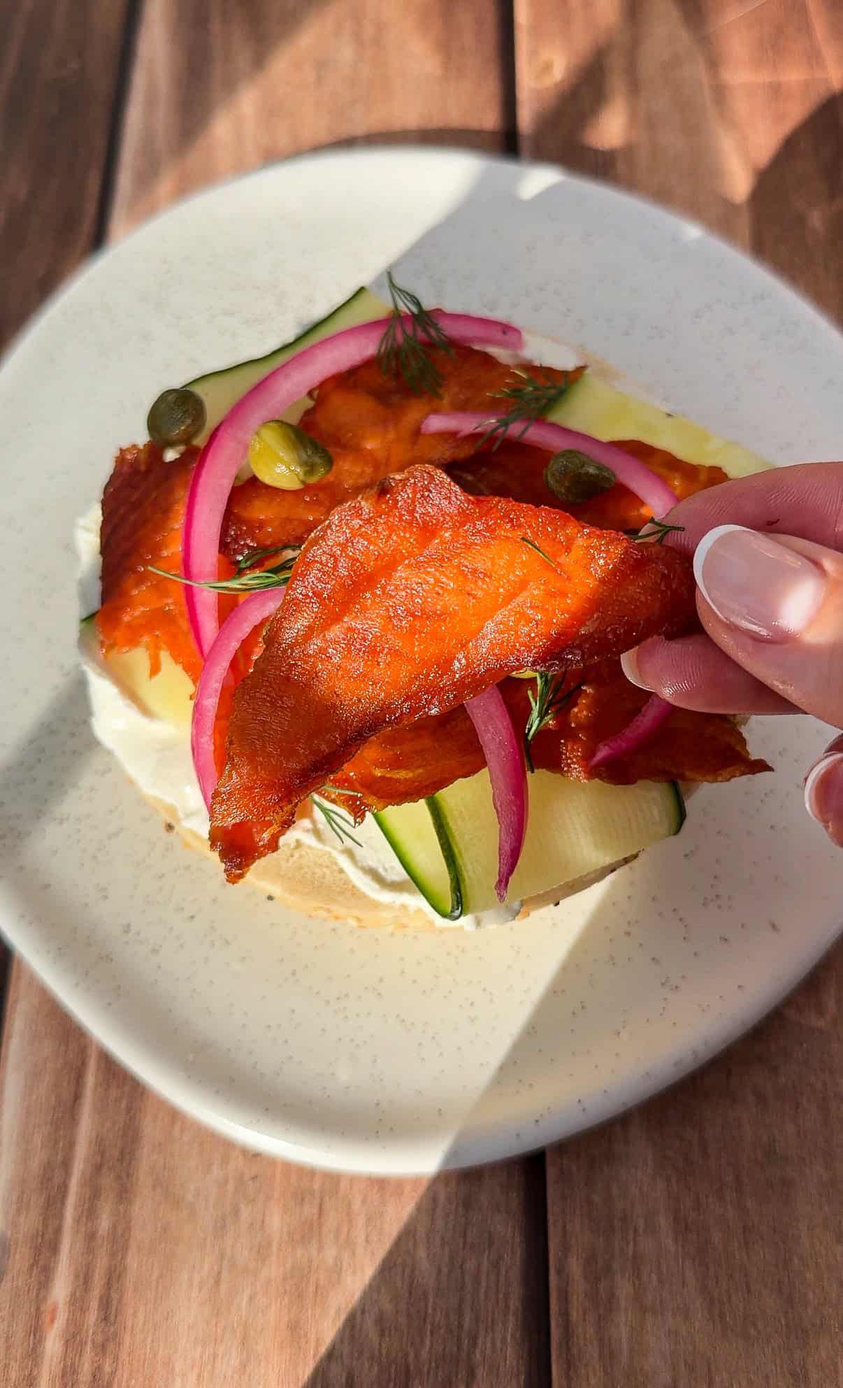 A hand holds a slice of plant-based smoked "salmon" above a bagel topped with cream cheese, zucchini ribbons, pickled onions, capers, and dill on a white plate, all on a wooden surface.