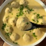 A bowl of creamy yellow soup with meatballs, garnished with chopped parsley. A wooden spoon is lifting one meatball from the bowl. The dish is set on a wooden surface.