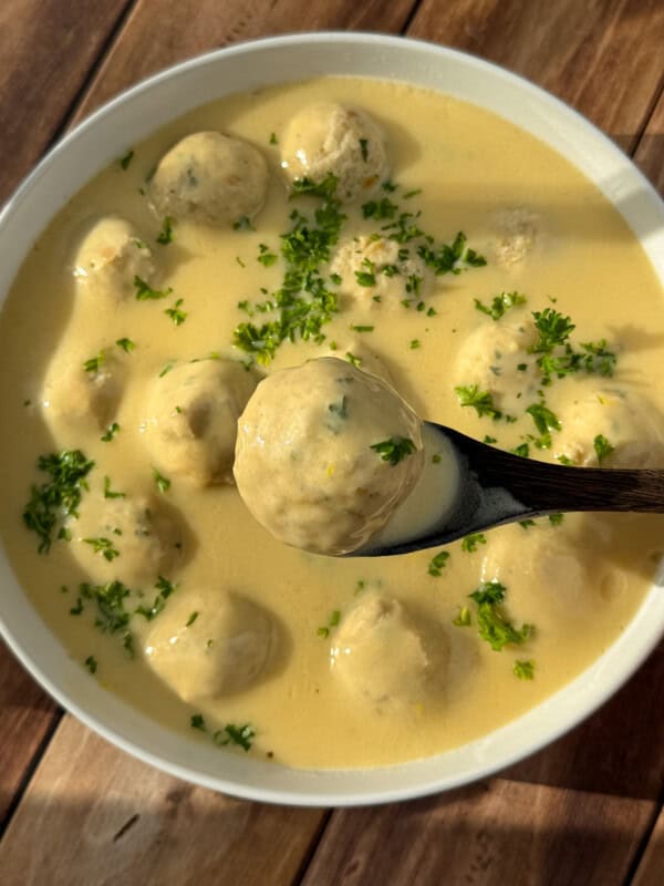 A bowl of creamy yellow soup with meatballs, garnished with chopped parsley. A wooden spoon is lifting one meatball from the bowl. The dish is set on a wooden surface.