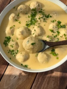 A bowl of creamy yellow soup with meatballs, garnished with chopped parsley, and a wooden spoon resting in the bowl. The dish is set on a wooden surface.