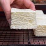 A hand holds a thick, square white marshmallow with a textured surface over a black wire cooling rack, with more marshmallows and a blurred background visible.