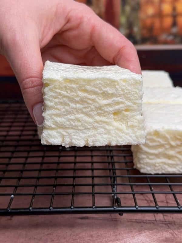 A hand holds a thick, square white marshmallow with a textured surface over a black wire cooling rack, with more marshmallows and a blurred background visible.