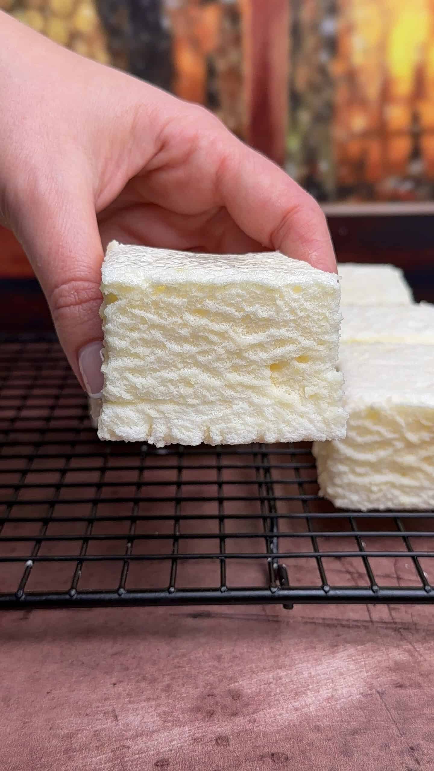 A hand holds a thick, square white marshmallow with a textured surface over a black wire cooling rack, with more marshmallows and a blurred background visible. - 4