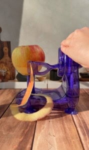 A hand turns the handle of a blue plastic apple peeler, peeling the skin off a red and yellow apple. The machine and apple sit on a wooden surface with sunlight streaming in.