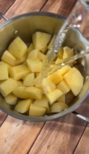 A pot filled with peeled, chopped potatoes is shown from above as water is being poured over them. The pot is placed on a wooden surface.
